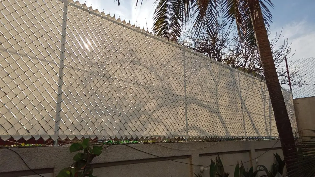 Malla ciclónica con cinta de privacidad beige sobre una barda, junto a una palmera